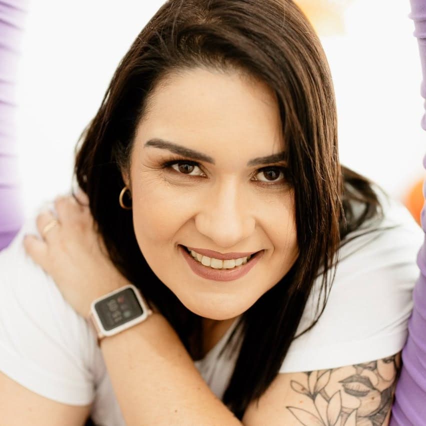 Carolina Almeida, terapeuta ocupacional, sorrindo entre dois tecidos de suspensão lilás usados em atividades de integração sensorial.